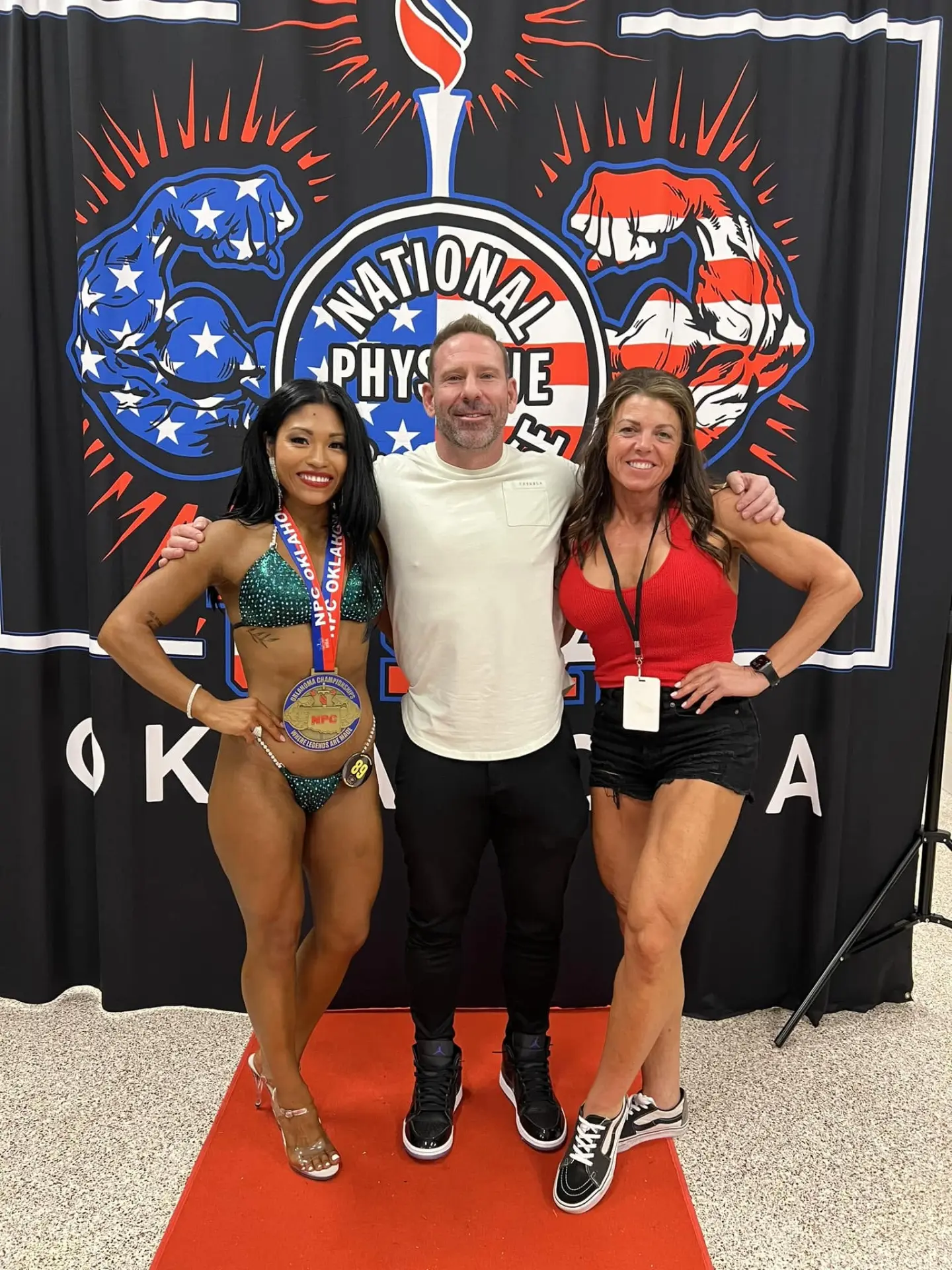 Ela Winder and Wendy Perdomo with their coach in front of the NPC Oklahoma Championship backdrop