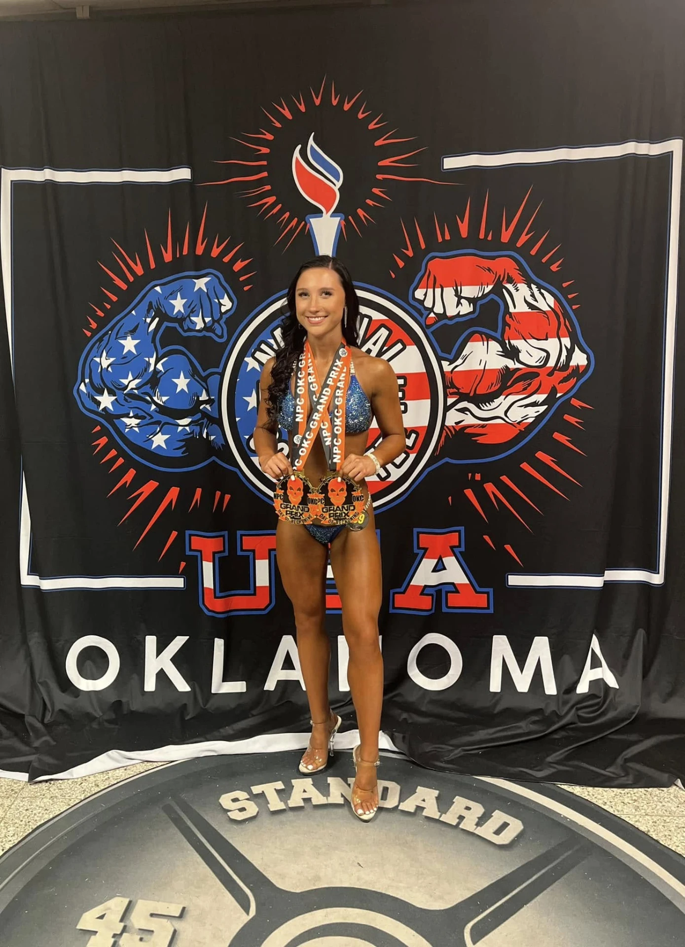 Jade White standing in front of the NPC Oklahoma backdrop holding her medals on competition day