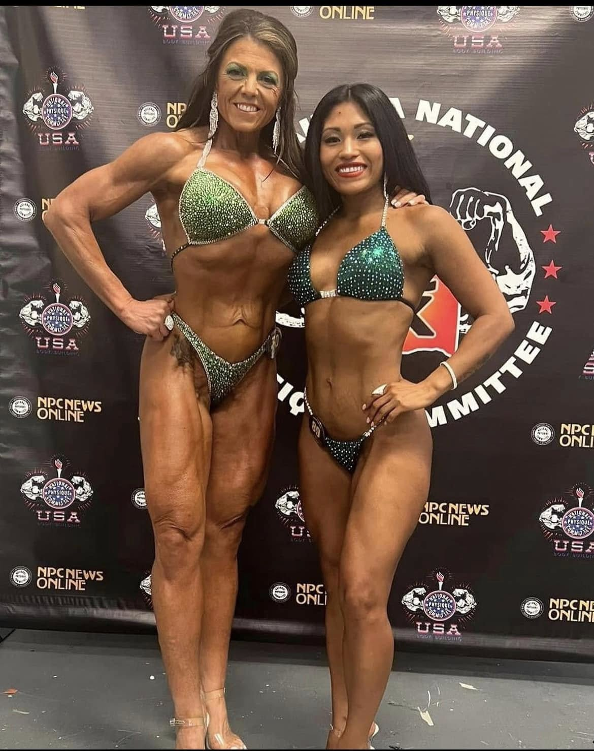 Two female NPC competitors in green suits posing together in front of a National Physique Committee backdrop