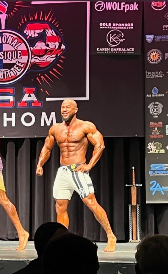 Men's physique competitor posing on an NPC Oklahoma stage in striped board shorts